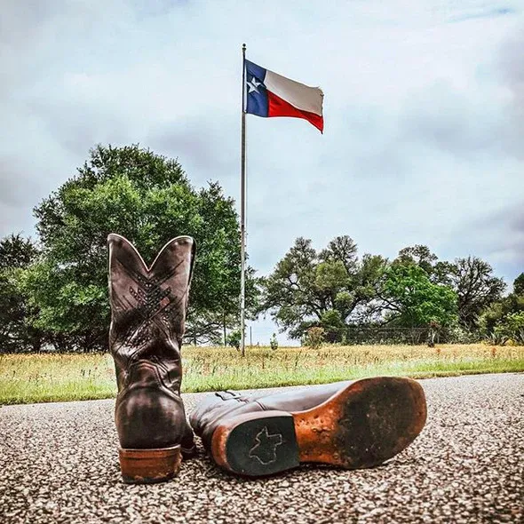 A pair of cowboy boots on a road, in front of a field with a Texas state flag.